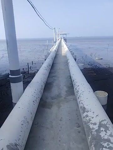 A first-person perspective video shows a walkway extending over shallow, muddy water under a clear sky. The walkway is flanked by two large, white pipes, likely for transporting water or materials. The walkway itself appears to be made of concrete. In the distance, a small structure with a roof is visible at the end of the walkway, possibly a lookout point or a pumping station. The surrounding environment is a vast expanse of water and mudflats, with some sparse vegetation or debris emerging from the water. The video is static, with no visible movement other than the slight wobble of the camera, suggesting it is handheld.
