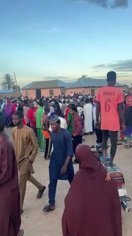 A crowd of people gathered outdoors under a cloudy sky. Many individuals are wearing traditional clothing, including tunics and head coverings. A person in a pink t-shirt with the number 6 on the back stands on a motorcycle, looking away from the camera. Others are standing and walking amongst each other. Buildings with light-colored walls and dark roofs are visible in the background. The scene appears to be a social gathering.