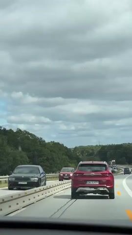 The video shows a view from inside a moving car on a highway. The sky is cloudy and gray. On the right side of the highway, there is a red SUV with a Polish license plate. On the left side of the highway, there is a red sedan. Further ahead, there is a construction zone with barriers, several vehicles including a white van and a construction machine, and a section of the road that appears to be under repair. There are also trees and foliage on both sides of the highway. The camera is panning slightly to the left.