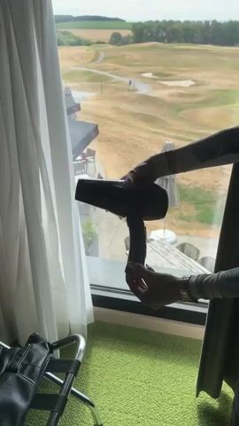 The video shows a person standing indoors in front of a large window. The person is holding a black hairdryer with a red cord in their hands. The person's hands are visible as they manipulate the hairdryer. The hairdryer is positioned in front of the window, and the person's arms are extended. Outside the window, a golf course is visible with rolling green hills and a clear sky. A luggage rack is partially visible on the left side of the frame. The floor inside is covered with a bright green carpet. The person is wearing long-sleeved clothing with a patterned design. The person is plugging the red cord of the hairdryer into a socket.