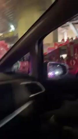 From inside a car, the video shows a scene with police motorcyclists moving through a crowd amid red smoke. The motorcycles are parked, with officers standing nearby. A bus with "SANTA FE" printed on its side is also visible. The camera is positioned inside the car, looking through the side window at the street scene. The lighting appears to be from streetlights and emergency vehicle lights. The overall environment suggests a potentially tense or chaotic situation.