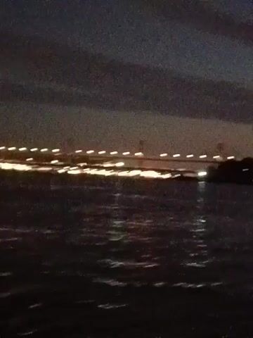 The video shows a night scene on the water. In the distance, a bridge is visible, lit up with numerous lights. The water is dark and rippling. The camera movement is unsteady, suggesting the footage was taken from a moving boat or another unstable platform. The overall atmosphere is dimly lit and the quality of the image is somewhat grainy. No people are visible.