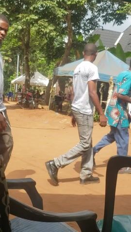 The video captures an outdoor gathering during the daytime in what appears to be a rural or semi-urban setting in Umundugba, Nigeria. The ground is unpaved, consisting of reddish-brown dirt or sand, and the weather is sunny with clear skies.

In the foreground, a man wearing a grey t-shirt and dark trousers stands, looking to his right. His arms are muscular, and he has a bracelet on his left wrist. Black plastic chairs are visible nearby. The camera then pans down and around, revealing the laps of the person filming, adorned with patterned fabrics; one has "SQUARE" and "PLUS" visible, while another features a chain-link design.

The background features lush green trees and foliage, suggesting a natural environment. A simple, light-colored building with a grey corrugated metal roof can be seen to the left. A light blue canopy tent is set up further back, under which several people are gathered, some seated. In the distance, a black car is parked, with what appears to be a speaker setup or a poster affixed to its rear, displaying partially legible text including "DJ KING" and "UMUD...". Background music plays throughout the video, indicating a lively atmosphere. The scene suggests a community event, possibly a celebration or social gathering.