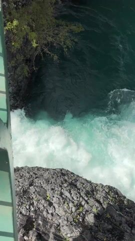 The camera films from above the churning, turquoise water of a powerful waterfall. The water foams and crashes against dark, jagged rocks. A green metal bridge edge appears in the upper left.