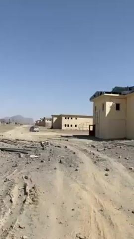 The video shows a line of two-story, light-yellow buildings, likely in a desert or arid environment. The buildings appear to be abandoned or in disrepair, with missing windows, damage to the roofs, and debris scattered around the site. The ground is sandy and rocky, with a dirt road leading towards the buildings. A white vehicle is visible in the distance. The sky is clear and blue, suggesting a sunny day. There is no observable human activity.