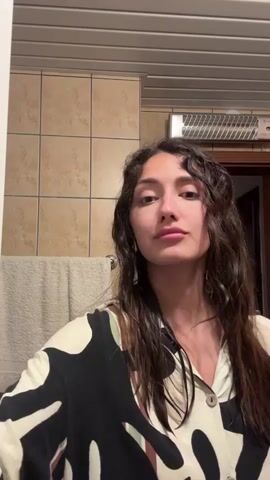 The video shows a young woman standing in a bathroom. She has long, dark, wavy hair that appears wet, and she is wearing a button-down shirt with a black and white abstract pattern. The bathroom has beige tiles on the wall, a towel hanging on a rack, and a ventilation fan near the ceiling. The woman looks straight ahead, then closes her eyes, then looks to the side before looking back at the camera. She sprays something into the air from a spray bottle with a blue nozzle.