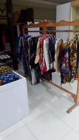The video shows the interior of a retail store that sells clothing and fabric. In the foreground, there is a wooden garment rack filled with colorful shirts and dresses. To the left, a white rectangular appliance, possibly a washing machine or storage unit, is partially visible. Further back, a display case with glass doors contains folded fabrics with intricate patterns. To the right, more clothing racks are arranged with various garments, and a tall white shelf unit displays folded fabrics in different colors and designs. In the background, additional clothing racks are visible, along with wooden display cabinets and shelving. The store appears to be well-lit and organized, with tiled flooring throughout. A glimpse of two individuals can be seen in the far background, suggesting the store may be open for business or staffed. The overall impression is of a neat and stocked boutique.