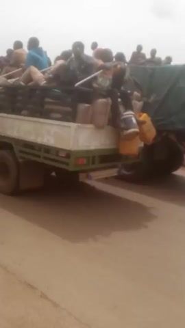The video shows a large truck with a green tarp-covered load bed.  Numerous people are riding on top of the tarp. The truck has multiple axles and tires.  The road is paved but dusty or sandy, with a reddish-brown color.  A motorcycle and a yellow auto-rickshaw pass by the truck. The sky is overcast and the light is flat.  The scene appears to be in a rural or less developed area.