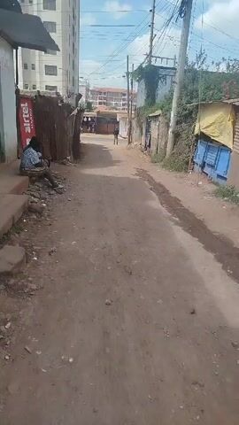 The video shows a dirt alleyway in what appears to be a lower-income area. A tall building is visible in the background. On the left side of the alley, two women are sitting on a concrete step. A large Airtel advertisement is behind them, partially obscured by corrugated metal. A dog lies on the ground nearby. On the right side of the alley, there is a corrugated metal structure covered with blue tarp. A man walks away from the camera down the alley. Debris is scattered along the sides of the alley. The sky is bright blue with some clouds.
