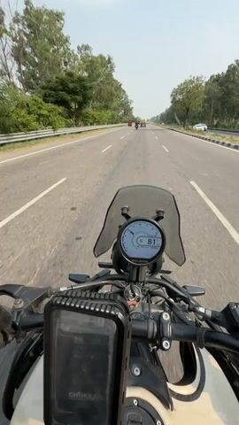 This is a first-person perspective video of someone riding a motorcycle on a highway near Nilokheri, India. The rider is wearing a red and black helmet, blue mirrored sunglasses, and a black motorcycle jacket with mesh accents. They are also wearing black gloves. Attached to the handlebars are prayer flags, displaying multiple colors and symbols.

The motorcycle has a digital display showing a speed of 81. A black phone mount or storage box is also attached to the handlebars. The highway is paved and appears to be a multi-lane road with a shoulder. Trees and vegetation line the sides of the highway, with some taller trees visible in the distance. The sky is bright and appears to be a clear or partly cloudy day.

Several other vehicles are visible on the highway, including cars and trucks. The overall scene suggests a daytime journey on a road with moderate traffic. The rider's perspective, combined with the movement of the prayer flags, conveys a sense of travel and potentially a spiritual element due to the flags. No explicit emotions are conveyed, but the activity itself suggests enjoyment or purpose in the ride.

The text "CHOKKA" is visible on the black storage box attached to the handlebars. The rider's t-shirt has text in a Cyrillic alphabet, though it is partially obscured and difficult to read clearly.