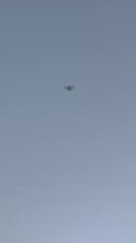 The video displays a drone flying in a clear, light blue sky. The drone is a small, dark object with four propellers, appearing as a cross shape. It is positioned in the upper center of the frame and appears to be stationary or moving slowly. No people or other objects are visible in the video.