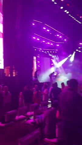 The video shows a large concert venue with a crowd of people gathered in a VIP section with tables and chairs. The stage is visible in the background with bright, purple lights and large screens displaying images. Some people are standing and mingling, while others are seated at tables. The lighting is dim, with the stage lights providing the primary source of illumination.