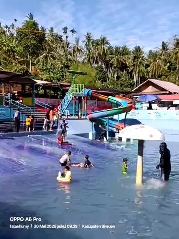 The video shows children playing in a water park on a sunny day. The water park features a large pool with a tiled floor, a water slide, and a mushroom-shaped sprinkler. Several children are visible in the pool, some are sitting or lying down, while others are walking around and splashing water. There are also adults and older children standing on the steps leading to the water slide. The background includes tropical trees and some buildings. The video quality is decent.