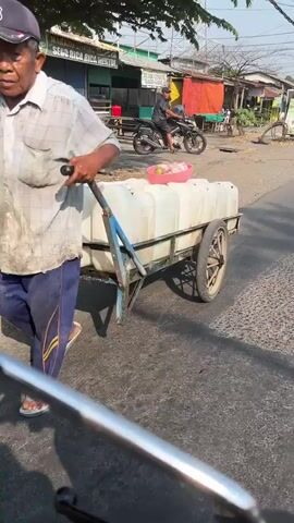 An elderly man with weathered skin and a blue baseball cap is walking on a paved road. He is pushing a cart with a white tarp and two large white containers. The cart has two wheels and a blue handle. The man is wearing a light-colored plaid shirt and dark pants with yellow stripes. He is also wearing sandals. In the background, there are buildings with signs in a foreign language, a motorcycle with a rider, and trees. The sun appears to be shining brightly, casting shadows. The overall scene depicts a person engaged in transporting goods on a street, likely in a commercial or residential area.