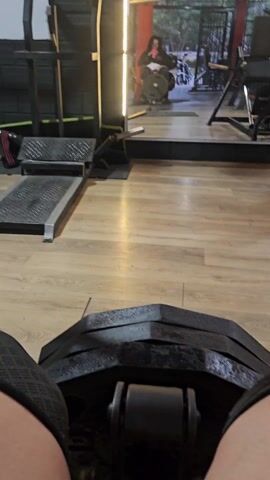 The video shows a first-person view from someone sitting on a leg press machine in a gym.  The weight plates are stacked on the machine in front of the viewer, and part of the person's legs and shorts are visible. The gym has wooden floors and various weight machines are visible.  A person is seen using a machine in the background near a mirror. The gym has red and white ceiling lights. The music is playing in the background.