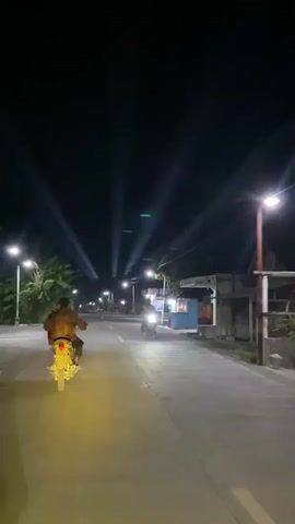 The video shows a nighttime scene on a paved road.  Multiple spotlights are angled upwards into the dark sky, creating distinct beams of light.  Several motorcycles are traveling along the road, illuminated by their headlights and the ambient light.  On the left side of the road, there are trees and some smaller structures. On the right side, there are more buildings, appearing to be shops or small establishments, some with lights on.  People are visible riding on the motorcycles, with one motorcycle in the foreground with two riders, one wearing a headscarf. The overall atmosphere is that of a road at night with some form of event or lighting display occurring.