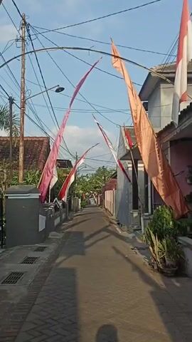 The video shows a narrow, paved alleyway lined with houses and fences. Several Indonesian flags, red and white, are strung across the alleyway and flutter in the wind. Power lines crisscross overhead. In the distance, a person on a motorcycle rides down the alley. The sun casts long shadows across the pavement, indicating it is either early morning or late afternoon. The scene appears to be a residential neighborhood.