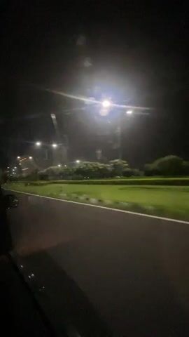 A car is driving on a road at night. The road is paved and has white lines marking the lanes. To the right of the road, there is a grassy area with some trees and bushes. There are streetlights illuminating the area. The sky is dark, and there are some lights visible in the distance, possibly from buildings or other vehicles. The video is taken from inside the car, looking out the window. The camera is slightly shaky, indicating that the car is in motion.