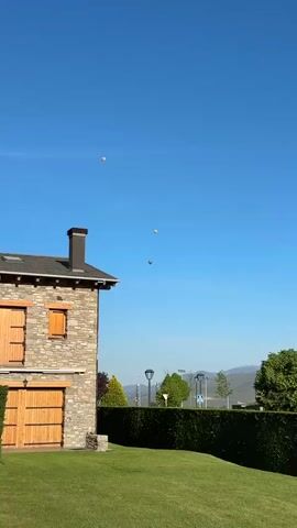 The video shows three hot air balloons rising in a clear, blue sky. The balloons are different colors: one is yellow, one is patterned, and one is gray. The camera pans up and follows the balloons as they rise. In the foreground, there's a stone building with a dark gray roof and a tall chimney. There's also a neat green hedge and some trees. In the far distance, low mountains are visible. The sky is mostly clear, except for the balloons and a thin white line that appears to be a contrail. There is some indistinct background noise and what sounds like indistinct speech.