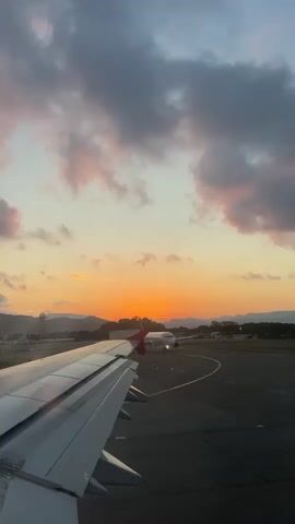 The video shows a view from an airplane window at an airport during sunset. The airplane wing is visible in the foreground on the left. Another airplane is seen on the runway in the middle ground, facing away from the camera. The setting sun, a bright orange disc, is visible above the horizon between the two airplanes. In the background, there are mountains and some trees. The sky is a mix of orange, yellow, and blue, with some clouds.