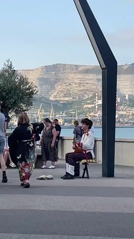 The media file captures a lively scene outdoors, likely on a promenade or waterfront, with a mountainous industrial landscape in the background. The time of day appears to be late afternoon or early evening, with the sky a soft blue and the sunlight casting long shadows.

In the foreground, a young person dressed in traditional clothing, including a white shirt with red trim and patterned trousers, sits on a chair playing a stringed instrument that resembles a lute or a balalaika. Their posture and engagement with the instrument suggest they are performing.

To the left, a woman in a flowing, colorful dress and a dark kimono-like garment is also in motion, possibly dancing or preparing to dance. Near her, a young girl in a white t-shirt and shorts sits on a ledge, observing the performance. Other individuals are visible in the mid-ground, some standing and watching, others seated, creating a relaxed and audience-like atmosphere.

The background reveals a harbor with cranes and industrial structures, including smokestacks, against a backdrop of layered, terraced mountains. The sea stretches out between the promenade and the industrial area. The overall mood is one of casual enjoyment and cultural engagement, with street performance adding a vibrant element to the setting. No visible text is present in the media file.