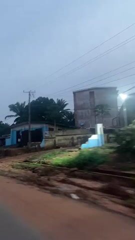 The video shows a street scene, possibly in a village or town. It is daytime, but the sky is overcast, suggesting it might be early morning or late afternoon, or possibly rainy. The road appears to be unpaved or partially paved, with red earth visible.

On the right side of the road, there are several buildings. One building is light blue with a dark roof and has a few people standing outside. In front of it, there are stacks of what appear to be bricks or building materials. Further down the road, there is a larger, multi-story building under construction, with scaffolding and exposed concrete. To the left of this building, there are some trees and other smaller structures.

There are several people visible in the video. Some are standing outside the blue building, and others appear to be working on the construction site of the larger building. There is also a motorcycle on the left side of the road in one of the frames. In the foreground, a person wearing a red garment is partially visible, suggesting the video is being filmed from a moving vehicle or from the perspective of someone within a vehicle. The movement of the camera is somewhat shaky, indicating it is likely handheld or mounted on a moving object. The overall impression is a glimpse of daily life in a developing area.