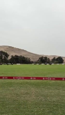 The video shows a polo match in progress on a green field. Several players on horseback are visible in the distance, actively engaged in the game. A red banner with the UBS logo repeats multiple times is visible in the foreground, bordering the field. The background features a tree line, followed by rolling hills and a cloudy sky. The overall atmosphere suggests an outdoor sporting event.