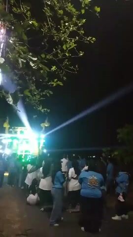 This is a short video clip shot at night, likely outdoors, in Dukuh Klopo, Indonesia. The main subject is a crowd of people gathered in front of a large sound system and stage, illuminated by a dynamic light show featuring bright beams of white, blue, and green lights. The people appear to be watching or participating in an event, possibly a concert or festival. Many are wearing casual clothing, and some are wearing blue shirts with a logo that is partially visible. The time of day is clearly night due to the dark sky and the prominence of the artificial lighting. The setting appears to be an open area, possibly a field or a street, with trees visible in the foreground. The atmosphere seems energetic and festive, driven by the music and lights. There are some flags or banners visible on the stage, and a sign above the speakers has some text, though it is difficult to read due to the lighting and resolution. The video is shaky, suggesting it was filmed handheld while moving.