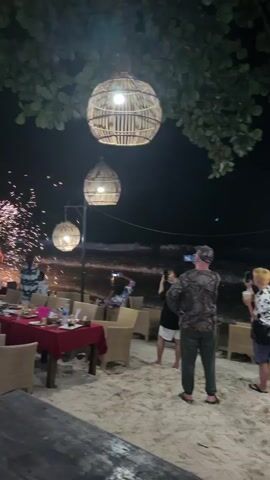 The video shows an evening beach scene with multiple people watching and recording a fire show. In the foreground, large woven basket-like lanterns hang from trees, illuminating a sandy area where tables and chairs are set up. One prominent table with a red tablecloth is visible, adorned with dishes and bottles. A man in a camouflage shirt and green pants stands in the mid-ground, facing the ocean and recording the activity with his phone. To his left, other individuals are also recording. Further left, a large, fan-shaped display of bright golden sparks erupts on the beach, drawing the attention of several people gathered around it. Waves are visible in the dark ocean background.