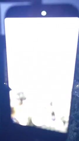 The video begins with a close-up of a smartphone screen in portrait mode, displaying a blurred image of a nighttime scene. The phone has a red recording indicator and a green status light at the top. The camera then pans slightly to the right, revealing the actual scene being captured. It shows a dark, possibly rainy, outdoor environment at night. In the foreground, out of focus, are what appear to be lights from the phone's camera or other nearby sources. The background features a building with illuminated windows and a row of parked cars on a wet surface, reflecting the lights from the building and streetlamps. Trees and darkness obscure the rest of the surroundings. The overall impression is of a nighttime recording of a residential area, possibly during or after rain.