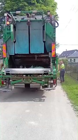 A garbage truck is parked on a paved surface. A man wearing a high-visibility vest and dark pants is standing to the right of the truck, facing it. He is holding a long object, possibly a tool or a part of the garbage truck's mechanism. The truck's rear is open, revealing the loading area. The man appears to be engaged in some activity with the truck. The background shows a residential area with houses and trees, indicating an outdoor, possibly suburban, setting. The lighting suggests it is daytime.