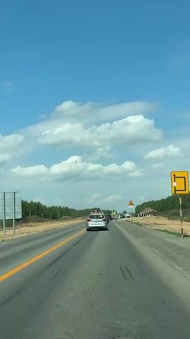 A car drives on a paved road under a cloudy sky. There is a yellow line on the left side of the road. The car is approaching a traffic zone with barricades on either side and a warning sign. There are trees on both sides of the road. Other cars are ahead. Music plays.