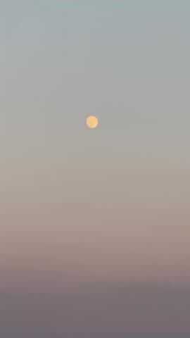 The video shows a distant view of a full or nearly full moon in a hazy sky, transitioning from a light blue at the top to pink and then a muted purple near the horizon. The moon is a pale yellow or orange orb. Below the sky is a calm body of water, likely the sea. To the left, a rugged, tree-covered cliff face juts out into the water. A portion of a dark structure, possibly a balcony or roof, is visible in the top left corner of the frame, indicating the viewpoint is from a high elevation. The scene appears to be at dusk or dawn, given the soft light and the visible moon. There are no people or animals visible in the frame, and no discernible actions are taking place beyond the subtle atmospheric color changes and the steady presence of the moon and landscape.