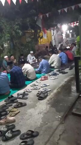 A group of men are gathered outdoors at night, sitting on the ground around food. They are facing each other and appear to be eating or socializing. There are many pairs of sandals and flip-flops neatly arranged on the ground in front of them. Red and white bunting is strung overhead, suggesting a celebration or special occasion. The lighting appears to be artificial, possibly from lamps or house lights. The environment looks like a residential area, with trees and buildings visible in the background. The men are dressed in casual clothing, and some are wearing skullcaps, indicating a religious or cultural gathering.