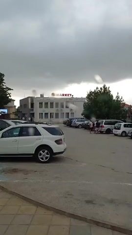 The video shows a parking lot with several cars. A white SUV is parked in the foreground. In the background, there is a building with a sign that says "MARKET". A group of people is gathered near a store in the parking lot. The sky is overcast and it appears to be raining.