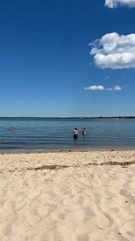 This is a vertical video clip of a tranquil beach scene. The foreground is dominated by a sandy beach, with some dried seaweed and debris scattered across it. Beyond the beach, a calm, blue body of water stretches towards the horizon. The sky is a vibrant, clear blue, with a few fluffy white clouds scattered across it. The time appears to be daytime, likely late morning or early afternoon, given the bright sunlight.

In the water, closer to the shore, three individuals are visible. Two of them, a male and female, are standing waist-deep in the water, facing away from the camera. Another person is partially submerged further out. In the distance, a sailboat with its white sail up is gliding across the water, adding a sense of gentle movement to the scene. The overall atmosphere is one of peace and relaxation, suggesting a pleasant day at the beach. No text is visible in the video. The location is identified as Flanders, United States of America.
