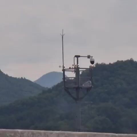 A weather monitoring station stands tall against a backdrop of lush green mountains and a cloudy sky. The station is equipped with various instruments including a wind vane, anemometer, and a camera, suggesting it is used for meteorological data collection. The camera, positioned at the top of the station, offers a panoramic view of the surrounding landscape. The scene suggests a remote location, possibly in a mountainous region, where such monitoring stations are often deployed to gather weather information. The overall atmosphere is serene and natural, with the weather station standing as a testament to human technological presence in the wild.