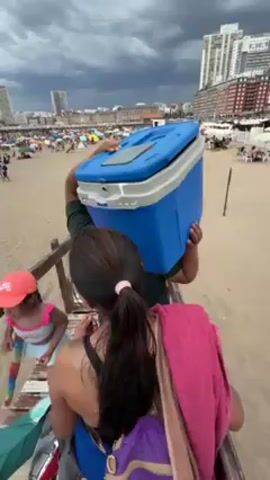 The video shows a person walking down a wooden staircase towards a crowded beach, carrying a large blue and white cooler on their head. Behind the person with the cooler, a woman with long dark hair in a ponytail carries a pink towel and a purple backpack. A child in a pink hat and blue and white swimsuit walks in front of them.  Other people are using the staircase, including an older woman wearing a hat and patterned jacket. The beach is filled with people, umbrellas, and tents.  In the background, tall buildings line the horizon under a cloudy sky.