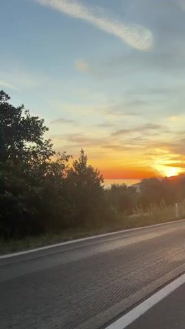 The video captures a scenic view of a sunset over a body of water, likely the sea, viewed from a moving vehicle on a paved road. The sky is a gradient of blue and orange, with wispy clouds. The sun is setting on the right side of the frame, casting a warm glow. Lush green trees and vegetation line the left side of the road. The road itself is asphalt with a white stripe marking the lane. Several cars are visible on the road, indicating traffic. The camera appears to be positioned inside a vehicle, possibly looking out the passenger window or from a dashcam. The overall impression is one of a peaceful drive during a beautiful sunset.