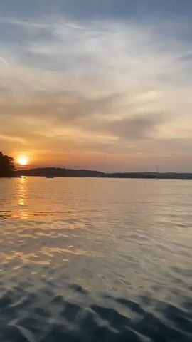 This is a vertical, wide-angle, video of a body of water with a sunset in the background. The water is calm with gentle ripples. A dark silhouette of trees and a shoreline is on the left side of the frame. The sun is low in the sky, casting a golden orange glow on the water and clouds. There are a few boats in the distance on the water. The sky is a mix of blue, orange, and yellow hues.