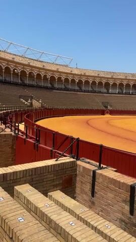 The media file displays an empty bullring in Seville, Spain, on a clear, sunny day. The sandy arena, a vibrant orange hue, occupies the center of the frame, bordered by a red wooden barrier. Above the barrier, tiered seating with an arched colonnade stretches around the arena, bathed in bright sunlight. The architecture suggests a historical and traditional venue.

The scene is devoid of any people or activity, creating a sense of stillness and anticipation. The bullring appears to be awaiting an event. Visible text includes seat numbers, such as "2" and "4," on the brick steps in the foreground and small numbered squares on the seats in the background. The bright, clear sky indicates daytime, likely midday given the intensity of the sunlight and the lack of shadows. The overall impression is one of grandeur and historical significance, with the emptiness amplifying the potential drama of a bullfight.