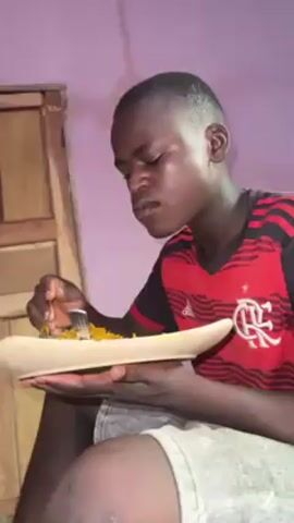 A young man is sitting down, holding a long, shallow dish filled with rice and using a fork to eat. He is wearing a red and black soccer jersey and denim shorts. The background includes a wooden door and a light purple wall.