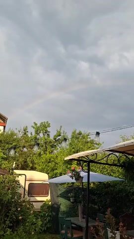 The video shows a view of an outdoor setting with a rainbow in the cloudy sky. In the foreground, there is a covered patio area with a table and chairs, and a string of lights overhead. To the left, a white caravan is partially visible. Lush green trees and bushes surround the scene.