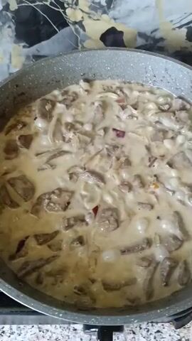 A close-up shot shows a gray non-stick skillet on a stovetop, filled with a creamy dish containing meat and mushroom slices. A pink silicone spatula is actively stirring the contents of the skillet. The background features a mottled countertop and a blurry backsplash with a gold and black abstract pattern. The lighting is natural, coming from above, and the overall scene suggests food preparation in a kitchen.
