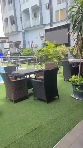The video captures an outdoor scene at what appears to be a hotel or resort in Oko Erin, Nigeria, during the daytime. In the foreground, there is an area with artificial green grass, furnished with several dark wicker-style outdoor tables and chairs, suggesting a dining or lounge space. A close-up shot briefly shows a black bag resting on one of these chairs.

Beyond the seating area, a bright blue swimming pool is visible, surrounded by a metal railing. On the far side of the pool, two multi-story buildings stand under a partly cloudy sky. The building on the left features modern architecture with large glass panels and balconies, while the building on the right has a more traditional design with smaller balconies. At least two individuals are seen near the far edge of the pool, one wearing a blue top and the other a white top, appearing to be walking or standing. Potted plants and air conditioning units are also visible on the premises, contributing to the serene, leisurely atmosphere.