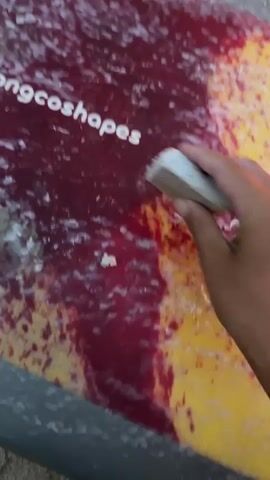 A hand is using a white object to scrub what appears to be a red and yellow object. The white object is creating white debris on the red and yellow surface. A coiled brown cord is visible in the background, along with sand.