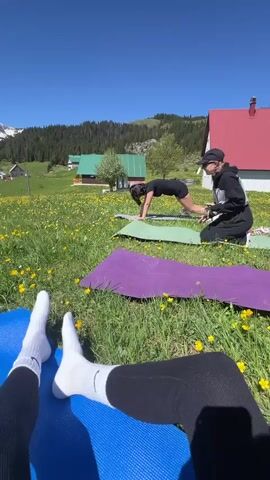 The video shows a first-person perspective of someone lying on a blue yoga mat in a grassy field with yellow wildflowers. Two other people are also on yoga mats in the background, with one person standing and adjusting her hair and the other kneeling down. In the distance, there are mountains partially covered in snow, interspersed with green forests. Small houses with pitched roofs are scattered across the landscape. The sky is clear and blue. The person recording the video is wearing black leggings and white socks.