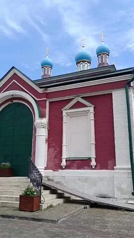 A slow-panning video captures the exterior of a religious complex, likely a monastery or church. The architecture is predominantly Russian Orthodox, characterized by red brick walls, white decorative trim, and multiple bulbous blue domes topped with gold crosses, visible against a cloudy sky. Several large, arched green doors are prominent features of the main building. The ground is paved with cobblestones, and there are stairs leading up to some of the entrances, with a ramp also visible for accessibility. Planter boxes with white flowers are placed near the entrances. The video also shows glimpses of adjacent buildings, including a long yellow structure with multiple windows and an arched gateway, as well as a smaller, older building with a brown, tent-like roof and a cross. The environment appears to be well-maintained and historic. There are no people or animals visible in the video.