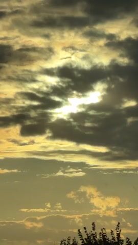 A time-lapse video of clouds at sunset, showing the sun peeking through the clouds, with the sky turning golden and yellow. The clouds are dark gray and white, and they are moving slowly across the sky. The sun is a bright white ball of light, and it is surrounded by a golden halo. The sky is a deep yellow, and it is filled with fluffy white clouds. The video is shot from a low angle, looking up at the sky.