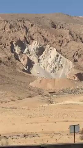 The video shows a desert landscape featuring a mountain with a large, light-colored quarry or mining operation on its side. A road with a few parked vehicles runs across the foreground. There's a single tree visible in the flat, sandy ground. The sky is clear and blue. The wind is loud.